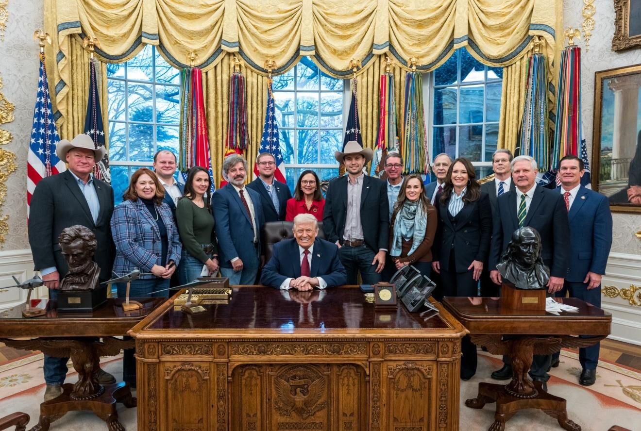 A group of people, several in cowboy hats and business attire, standing behind the desk in the Oval Office with the President seated at the desk.